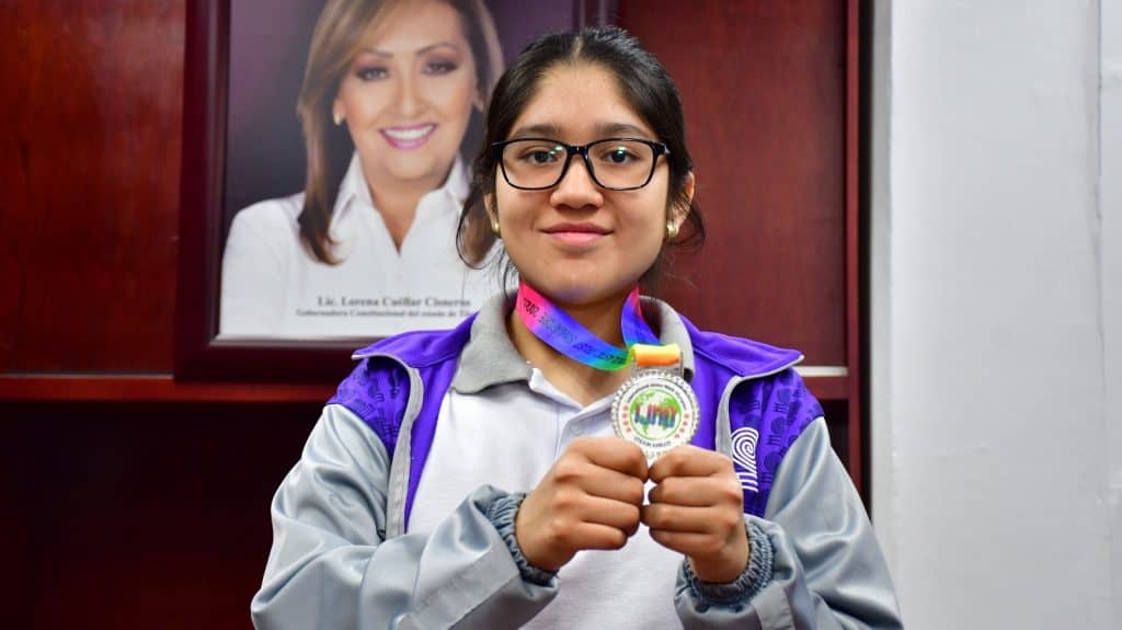 1. Niña con medalla de competencia de ciencia y tecnología, usando gafas y mochila, en interior con retrato de una mujer en el fondo.