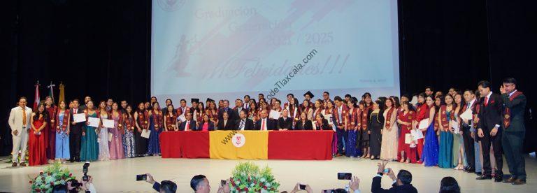 Estudiantes en ceremonia de graduación en un auditorio, con un escenario decorado y una pantalla grande que muestra un mensaje de felicitación por la graduación 2023.