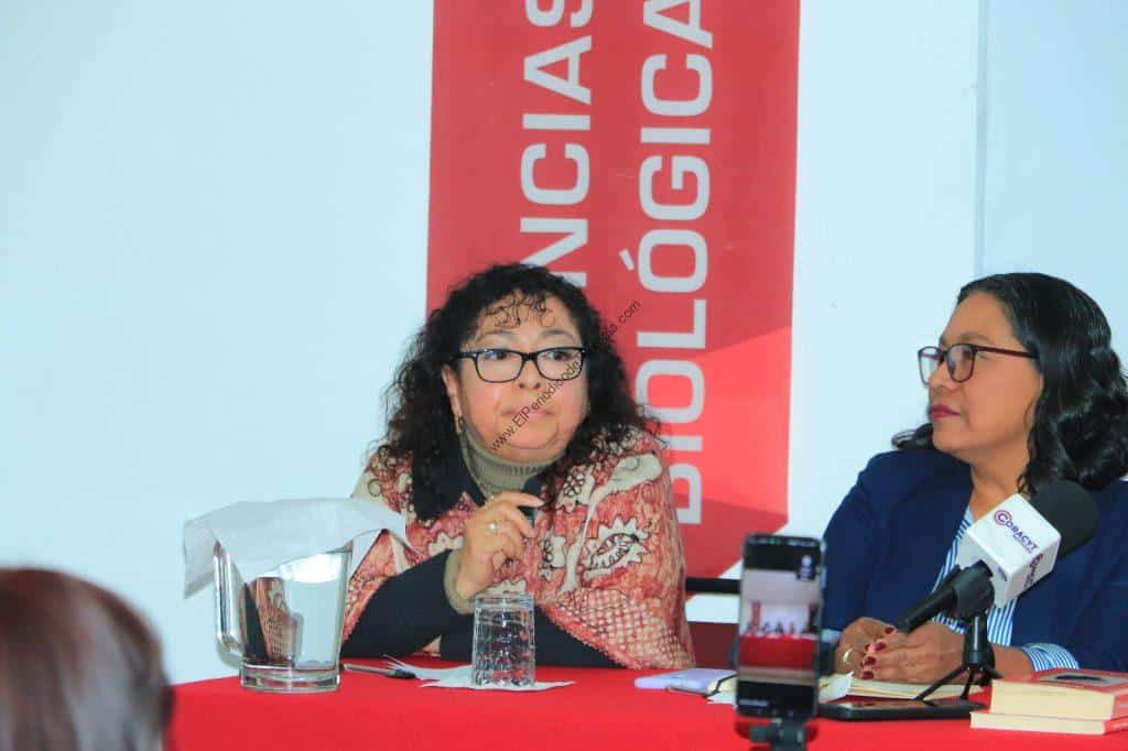 Dos mujeres en una mesa durante una conferencia, una de ellas hablando y la otra escuchando, con un cartel rojo de fondo que dice "Ciencias y Tecnologías".