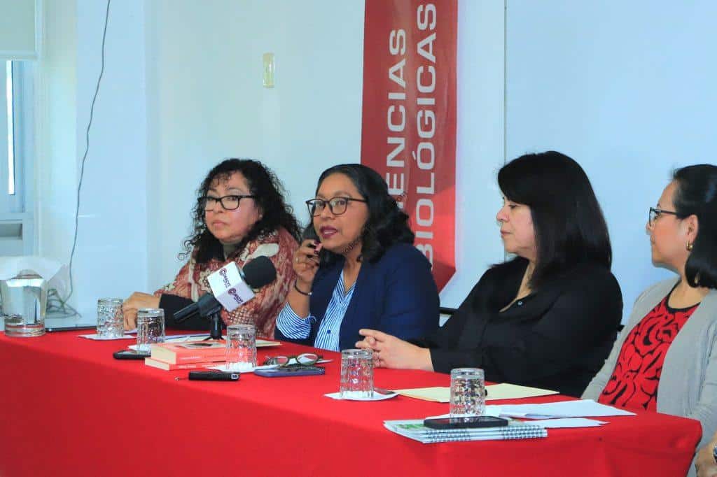 La mujer en el centro habla en una conferencia, rodeada de otras mujeres, en un evento formal sobre ciencias biológicas en Tlaxcala.