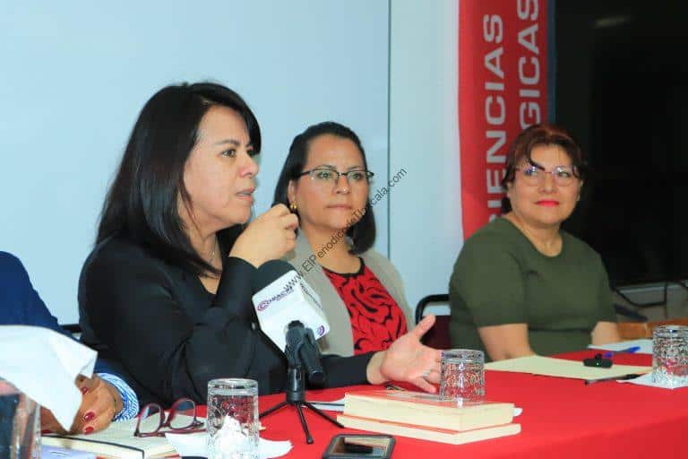 Mujeres en una mesa de discusión en un evento académico, con micrófono y documentos, participando en una conferencia o seminario en un entorno formal.