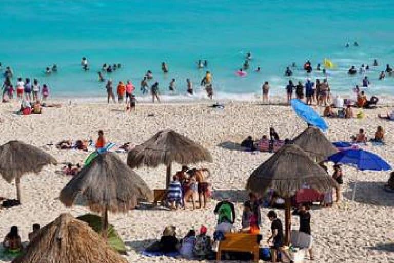 Disfruta de un día en la playa con muchas personas nadando y tomando el sol bajo sombrillas y palapas en un ambiente familiar y relajado.