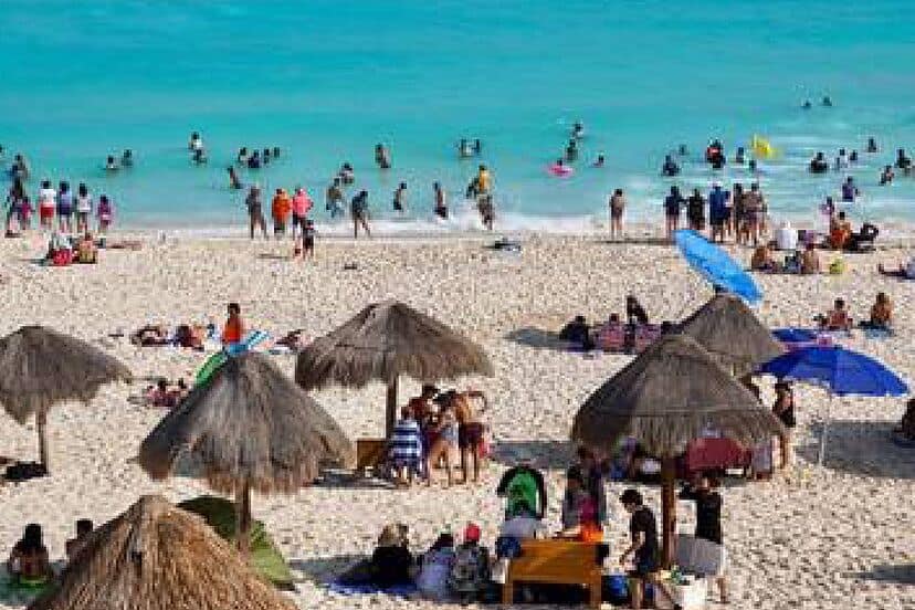 Disfruta de un día en la playa con muchas personas nadando y tomando el sol bajo sombrillas y palapas en un ambiente familiar y relajado.