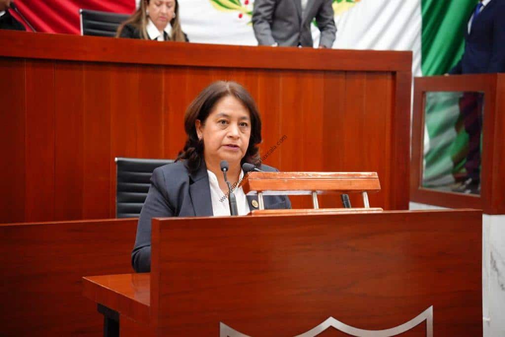 Mujer en un juicio o audiencia en un entorno formal, sentada en un escritorio de madera con micrófono, en un ambiente oficial y profesional.