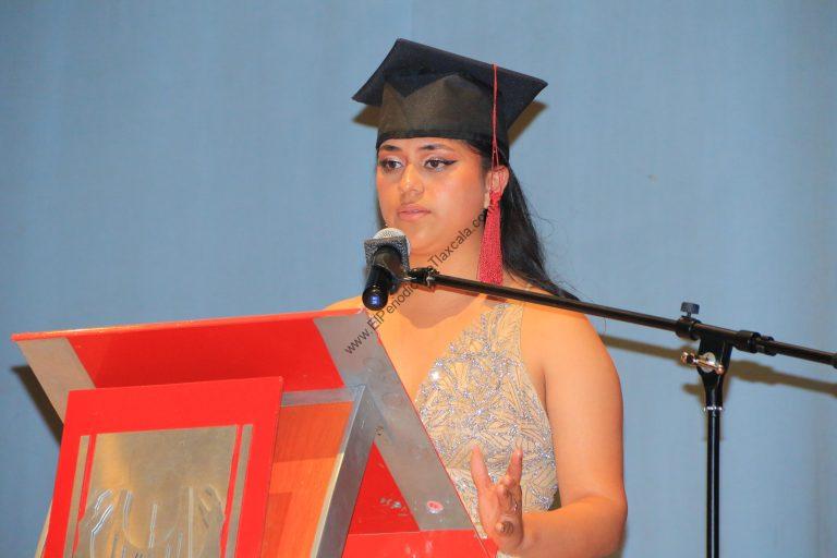 Estudiante con toga y birrete en ceremonia de graduación, hablando en un micrófono en un escenario con fondo azul.