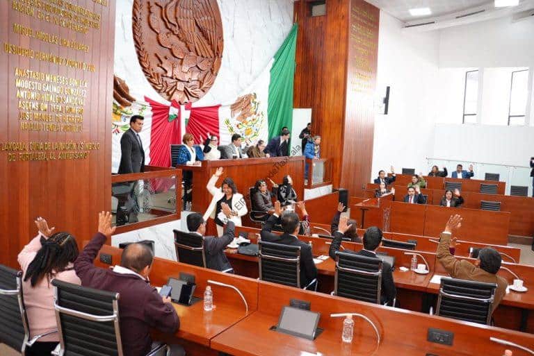 Miembros del Congreso de Tlaxcala en sesión, algunos levantando la mano, en un ambiente formal con banderas y escudo en el fondo.