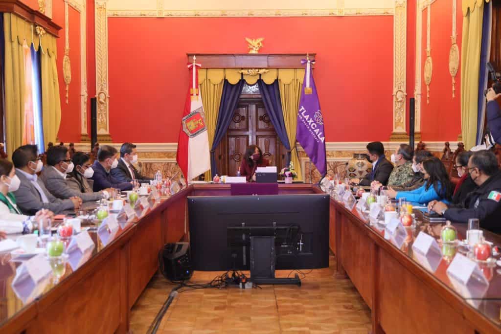 Reunión oficial en un salón con bandera de Tlaxcala y autoridades sentadas en una mesa larga, participando en un evento formal.