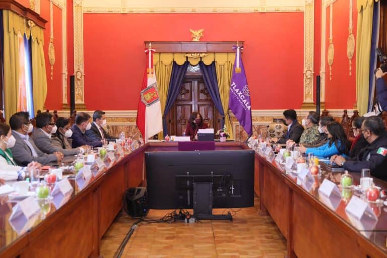 Reunión oficial en un salón con bandera de Tlaxcala y autoridades sentadas en una mesa larga, participando en un evento formal.