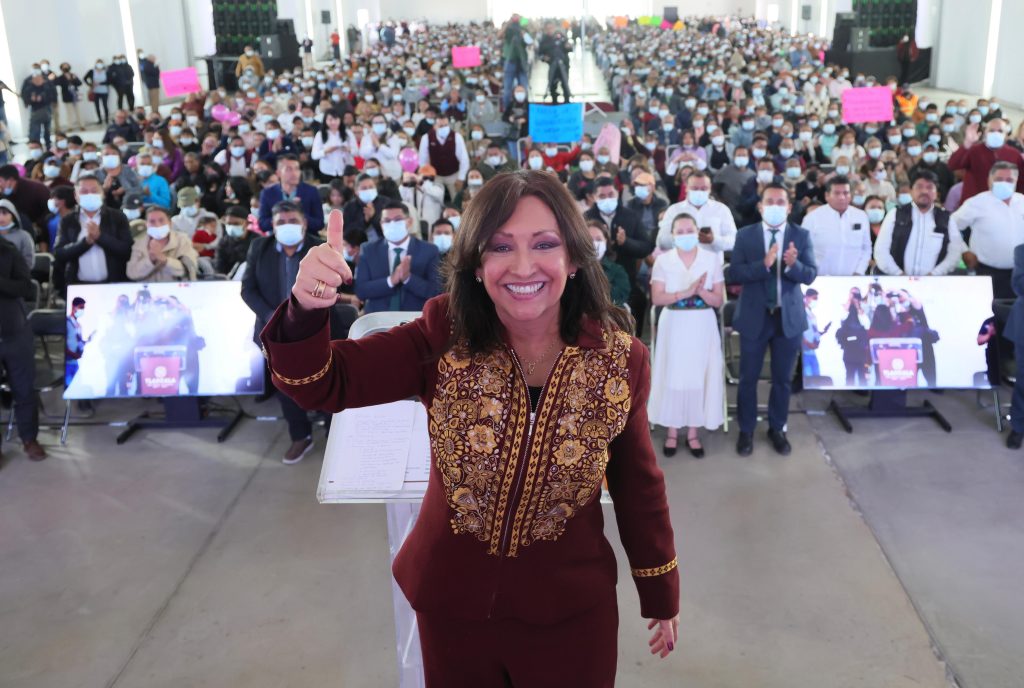 Mujer sonriente en evento masivo con multitud de personas usando mascarillas en un espacio cerrado.