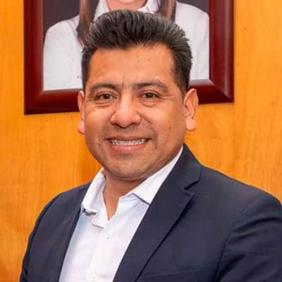 Hombre de negocios sonriendo, vestido con traje oscuro y camisa blanca, en un entorno profesional con fondo de madera y retrato enmarcado.