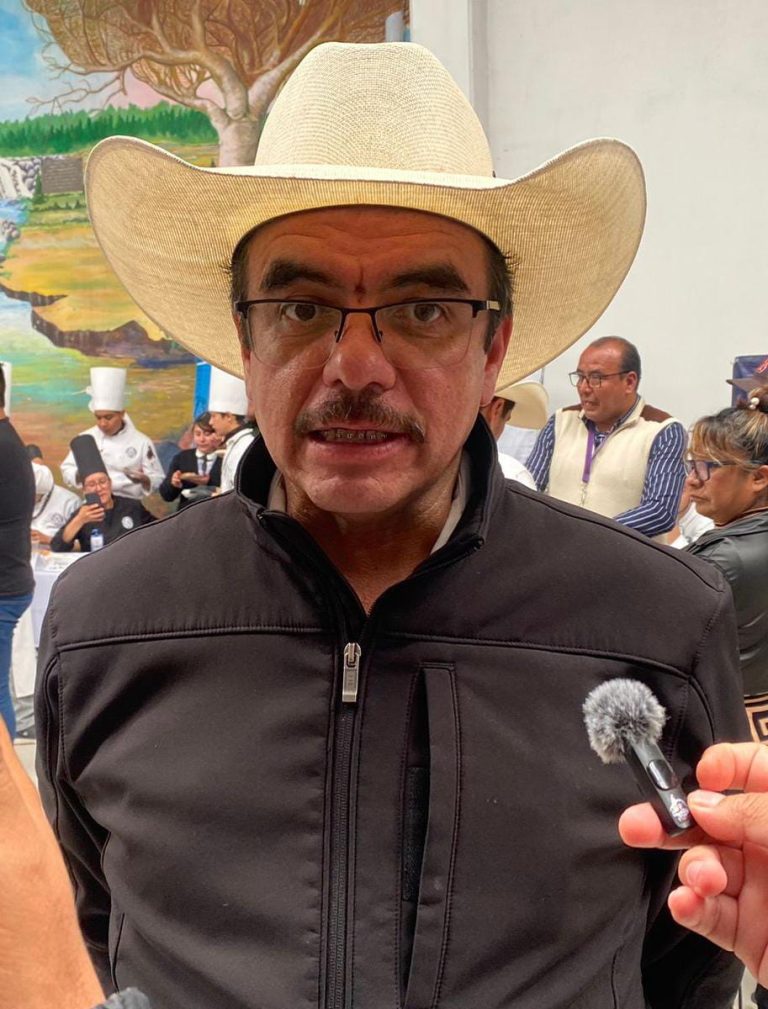 Hombre con sombrero de vaquero y gafas en una entrevista en un evento cultural, con varias personas en el fondo, algunas con trajes tradicionales y un mural en la pared.