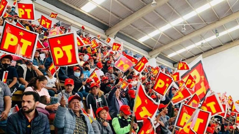Manifestantes con banderas rojas y amarillas del Partido del Trabajo (PT) en un evento político en un estadio cubierto, con asistentes de diferentes edades mostrando apoyo al partido.
