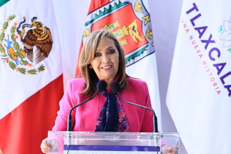Mujer en un podio con banderas de México, Tlaxcala y Tlalcala, hablando en un evento oficial, vistiendo un blazer rosa y una bufanda azul, en un entorno formal y político.