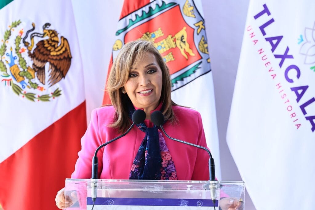 Mujer en un podio con banderas de México, Tlaxcala y Tlalcala, hablando en un evento oficial, vistiendo un blazer rosa y una bufanda azul, en un entorno formal y político.
