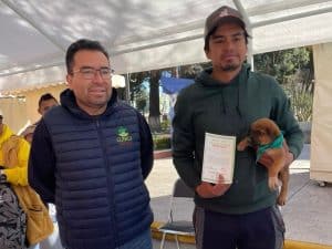 Dos hombres en evento al aire libre, uno con certificado y perro, en un ambiente con carpa y árboles.