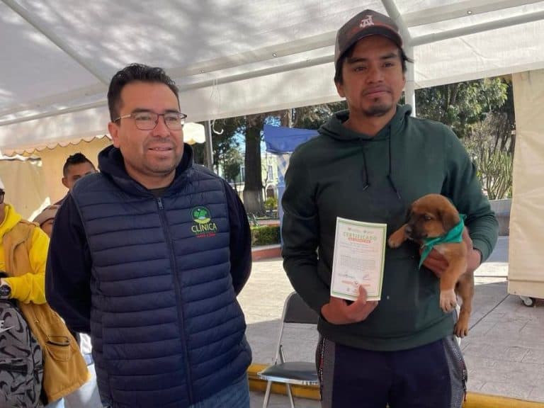 Dos hombres en evento al aire libre, uno con certificado y perro, en un ambiente con carpa y árboles.