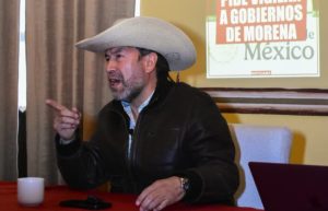 Hombre con sombrero y chaqueta negra hablando en una reunión, con cartel de fondo que promueve votar por gobiernos de Morena en México.