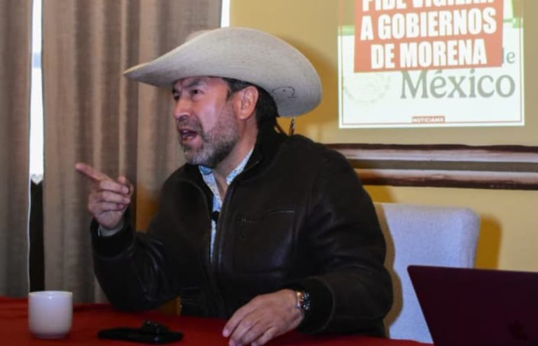 Hombre con sombrero y chaqueta negra hablando en una reunión, con cartel de fondo que promueve votar por gobiernos de Morena en México.