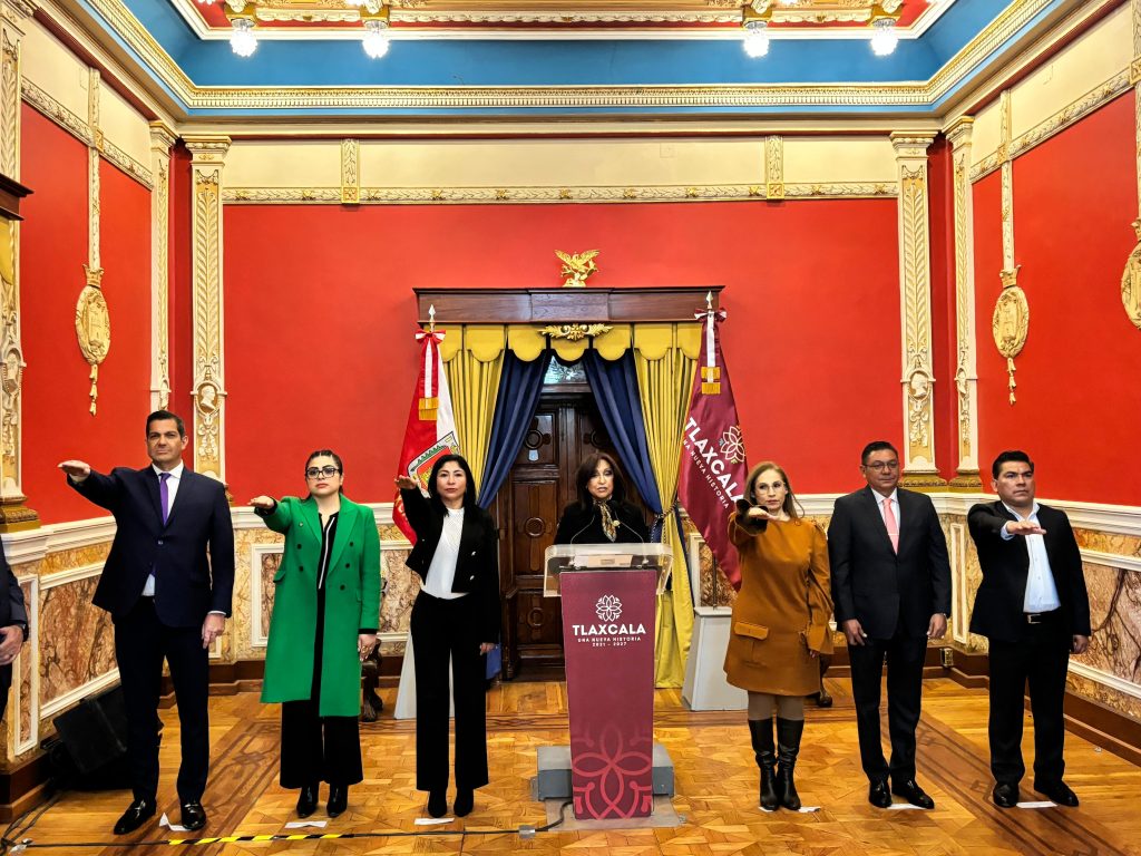 Grupo de personas en una sala decorada con colores rojo, dorado y azul, participando en un evento formal en Tlaxcala, México, con banderas y un podio en el centro.