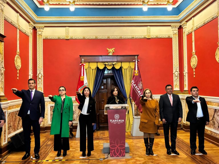 Grupo de personas en una sala decorada con colores rojo, dorado y azul, participando en un evento formal en Tlaxcala, México, con banderas y un podio en el centro.