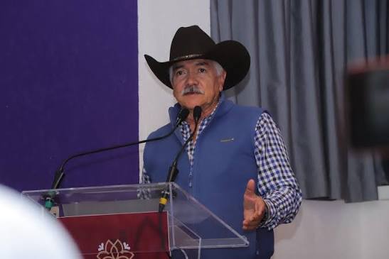 Hombre con sombrero cowboy, vestimenta azul y camisa a cuadros, hablando en un micrófono en un evento formal.