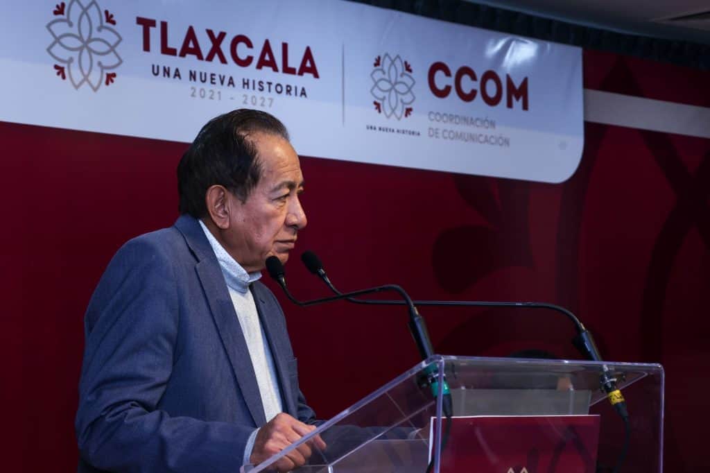 Hombre en traje azul hablando en un podio durante un evento oficial en Tlaxcala, con fondo de banner institucional y logotipos de la coordinación de comunicación.