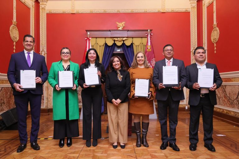 Grupo de seis personas en ceremonia formal, sosteniendo certificados en un salón decorado con paredes rojas y detalles dorados.