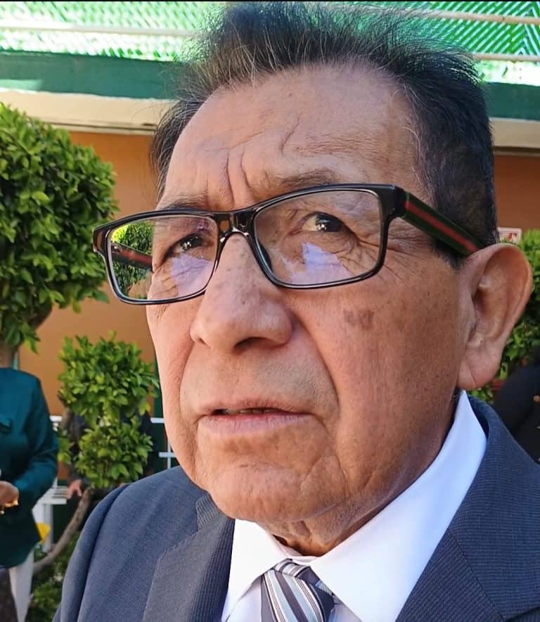 Retrato de un hombre mayor con gafas, vestido con traje y corbata, en un entorno exterior con plantas verdes y estructura de toldo.