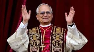 El Papa Francisco saludando con ambas manos en un acto oficial, vistiendo vestimenta religiosa tradicional y un fondo de cortinas rojas.