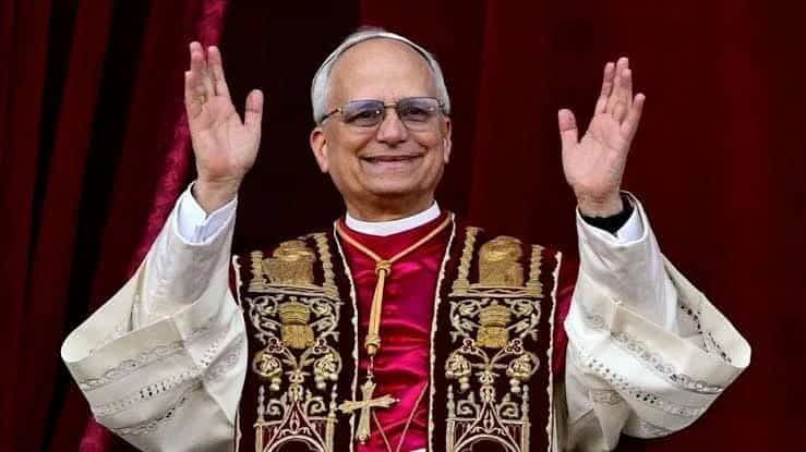 El Papa Francisco saludando con ambas manos en un acto oficial, vistiendo vestimenta religiosa tradicional y un fondo de cortinas rojas.