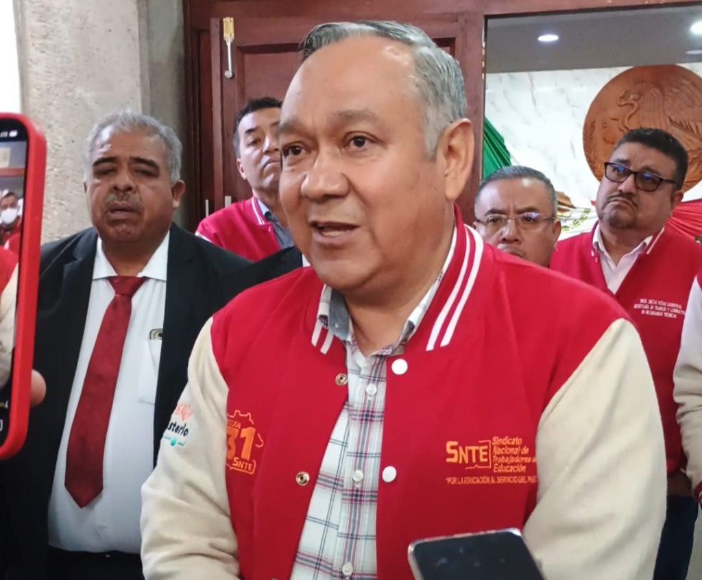 Hombre en una conferencia o evento, vestido con chaqueta roja con logotipos y texto relacionado con el sindicato SNT, rodeado de varias personas en un ambiente formal y de prensa.