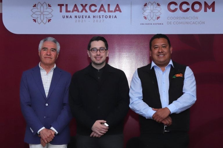 Una reunión formal con tres hombres de pie frente a un fondo con logotipos y texto relacionado con Tlaxcala y la coordinación de comunicación.