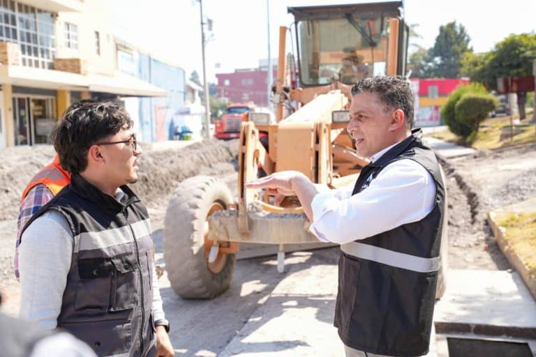 Dos hombres conversando en una obra de construcción en una calle de Tlaxcala, con maquinaria pesada al fondo y edificios en el entorno urbano.