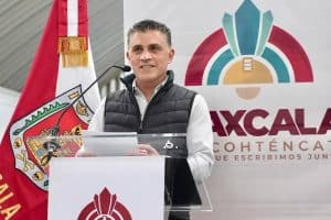 Hombre en podio durante evento oficial con bandera de Tlaxcala y logotipo en fondo, vestido con chaqueta negra y camisa blanca, hablando en público.