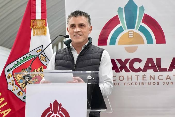 Hombre en podio durante evento oficial con bandera de Tlaxcala y logotipo en fondo, vestido con chaqueta negra y camisa blanca, hablando en público.