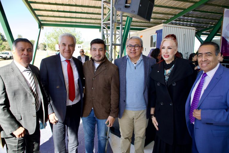 Grupo de seis personas en evento al aire libre, algunos con trajes formales, en un espacio con estructura metálica y fondo de árboles y cielo.