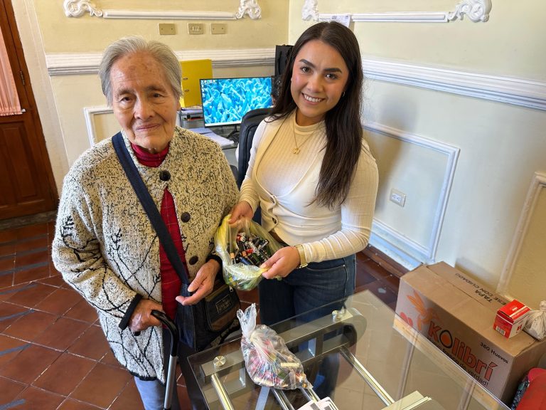Joven entregando un regalo a una mujer mayor en una oficina, en un ambiente interior con decoración clásica y tecnología moderna en el fondo.