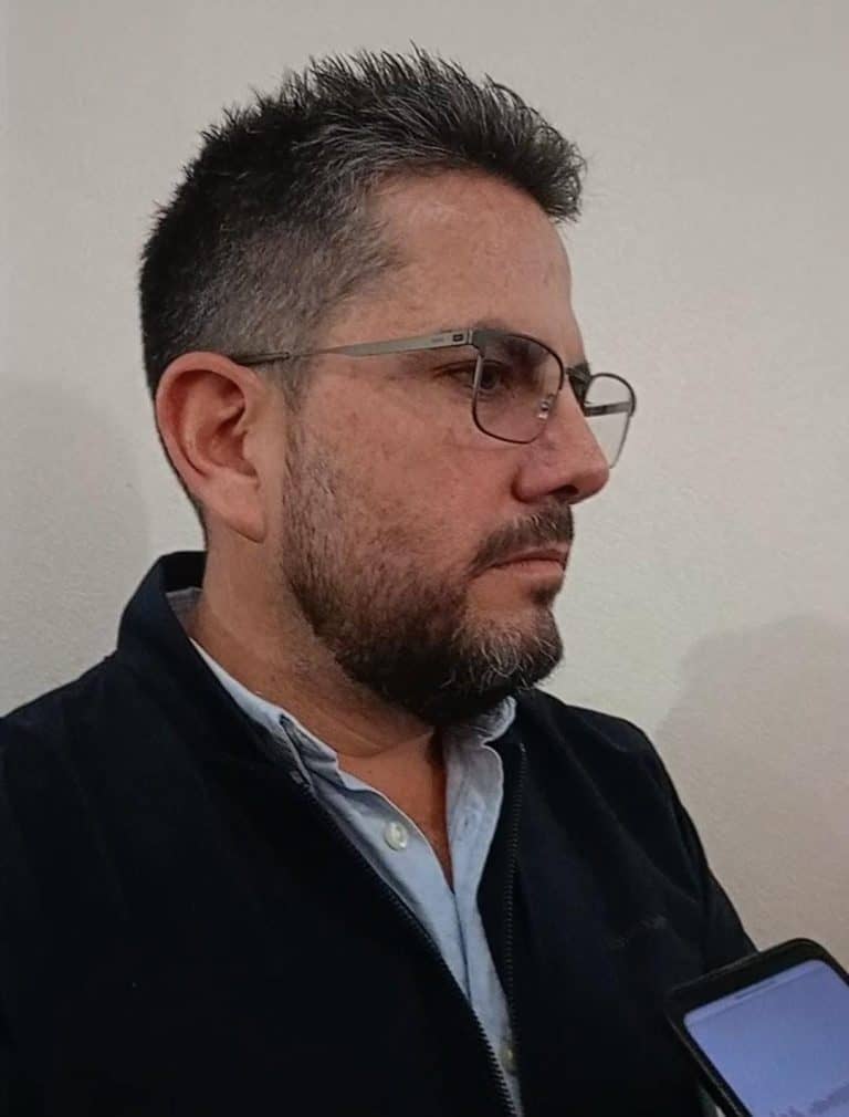 Hombre con barba y gafas mirando hacia la derecha, usando una chaqueta negra y camisa azul claro, en un fondo blanco.
