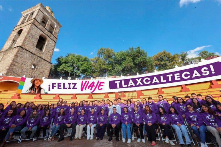 Grupo de personas con camisetas moradas en un evento al aire libre frente a una estructura histórica y un cartel que dice "¡Feliz viaje Tlaxcaltecas!".