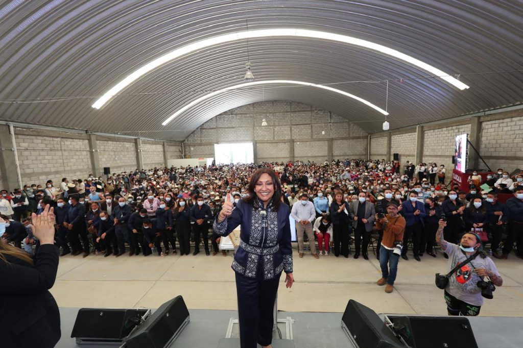 Mujer en el escenario con multitud de asistentes en un evento en un espacio cerrado con techo curvo y iluminación moderna.