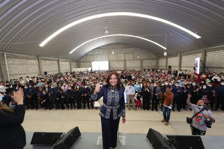 Mujer en el escenario con multitud de asistentes en un evento en un espacio cerrado con techo curvo y iluminación moderna.