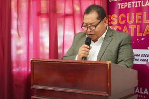 Persona hablando en un micrófono durante un evento en un escenario con fondo rosa y cartel de la escuela en Tlaxcala.