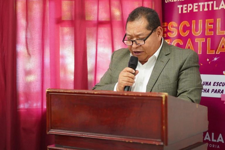 Persona hablando en un micrófono durante un evento en un escenario con fondo rosa y cartel de la escuela en Tlaxcala.