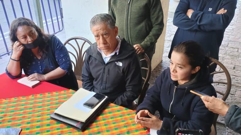 Grupo de personas en una reunión al aire libre, discutiendo temas importantes, con una mesa cubierta con un mantel colorido y varias personas observando atentamente.