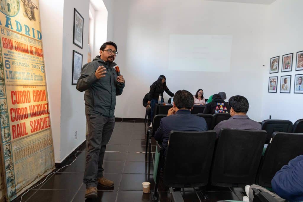 Hombre con gafas y chaqueta oscura dando una presentación en una sala de reuniones con asistentes sentados y una pantalla en la pared.