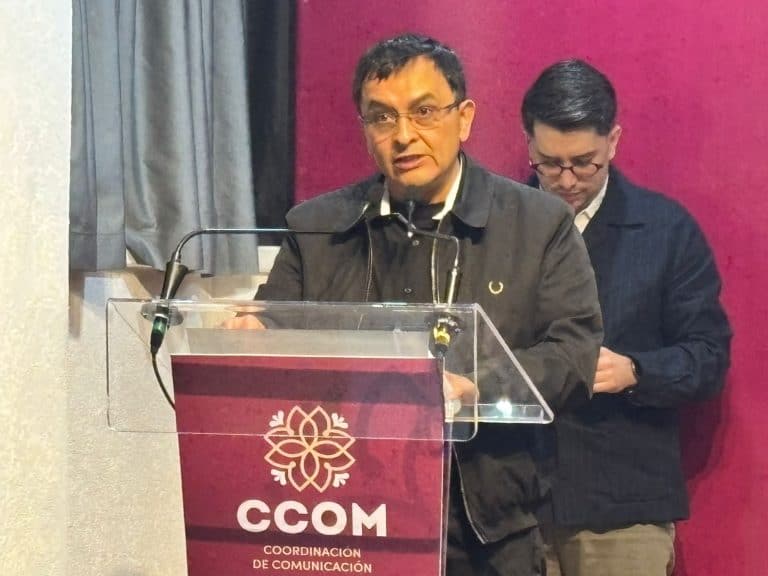 Hombre hablando en un podio con logotipo de CCOM en un evento formal, con fondo de cortinas grises y paredes rosas.