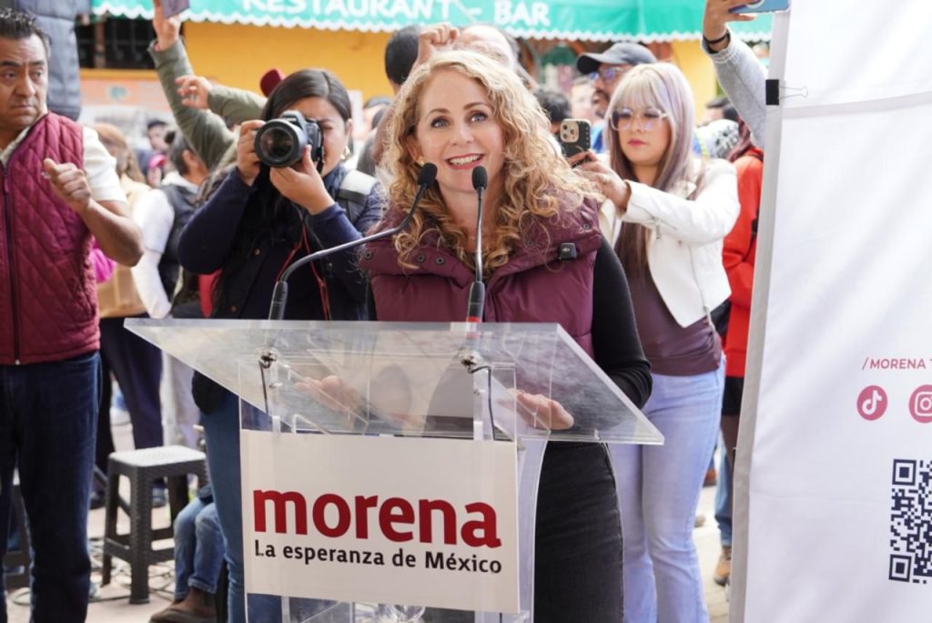 Una mujer sonriente da un discurso en un evento político, rodeada de varias personas que la apoyan y toman fotos, en un ambiente al aire libre con un cartel de "morena" y un banner de fondo.