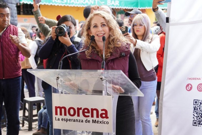 Una mujer sonriente da un discurso en un evento político, rodeada de varias personas que la apoyan y toman fotos, en un ambiente al aire libre con un cartel de "morena" y un banner de fondo.