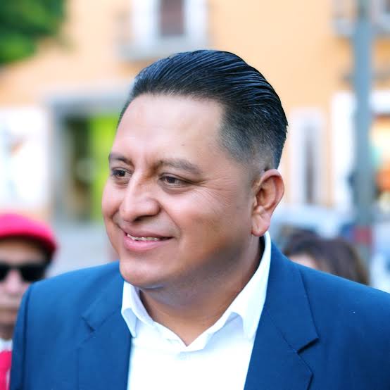 Hombre sonriendo con cabello peinado hacia atrás, vestido con traje azul y camisa blanca, en un evento público al aire libre en Tlaxcala, México.