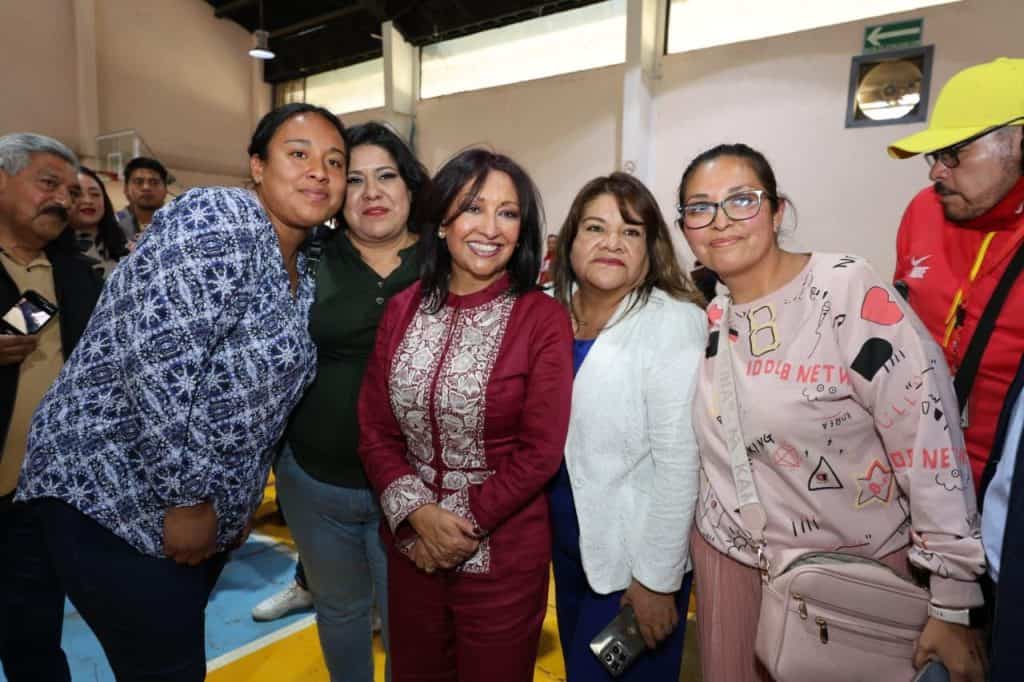 Mujer sonriente en grupo de personas en un evento público, con fondo de interior y varias personas observando.